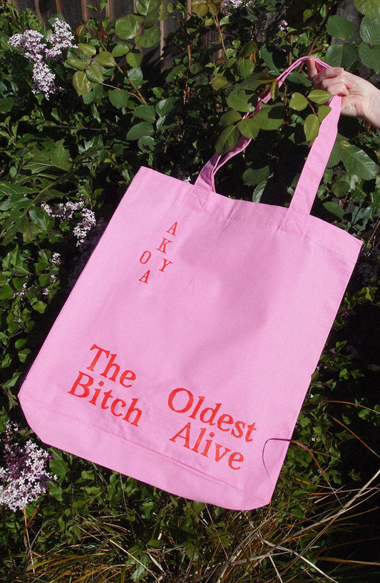 A pink canvas tote bag with red text reading 'The Bitch Oldest Alive' and 'AKY OA' printed vertically, held by a hand against a background of green foliage and small white flowers.